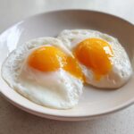 Close-up of two perfectly cooked poached eggs with bright, runny orange yolks served on a white plate.