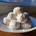 A stack of freshly baked russian tea cakes heavily coated in powdered sugar, sitting on a white plate near a window.