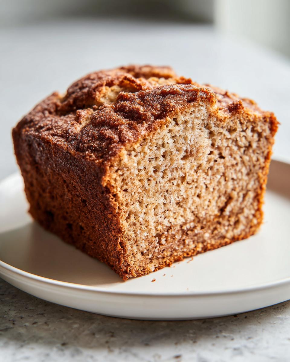 Close-up of a thick slice of moist Protein banana bread on a white plate, showing its textured crumb and cinnamon sugar top.