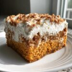 A square slice of pumpkin crunch dessert featuring a moist orange cake layer, thick white frosting, and crunchy pecan topping.
