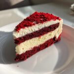 A perfect slice of red velvet cheesecake showing layers of deep red cake and creamy filling, topped with red crumbs.