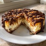 A whole Samoa Cheesecake with a slice removed, topped with toasted coconut, caramel, and chocolate drizzle.