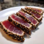 Five slices of perfectly seared ahi tuna, crusted with black and white sesame seeds, resting on a white plate.