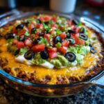 A close-up of a freshly prepared Seven layer dip topped with guacamole, sour cream, tomatoes, olives, and green onions.