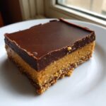 A single, rich square of peanut butter bars with a thick chocolate topping on a white plate.