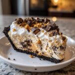 A generous slice of rich butterfinger pie with a dark crust, creamy filling, and topped with whipped cream and chopped candy.