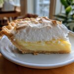A close-up of a slice of buttermilk pie featuring a thick layer of creamy filling and a tall, toasted meringue topping.