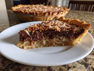 A close-up slice of german chocolate pie showing the coconut pecan layer and rich chocolate frosting, with the whole pie blurred in the background.