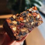 Close-up of a dense slice of fruit cake packed with dried fruits and nuts, held up to the light.