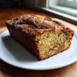 A freshly baked loaf of snickerdoodle banana bread with a sugary, cinnamon-swirled top, resting on a white plate.
