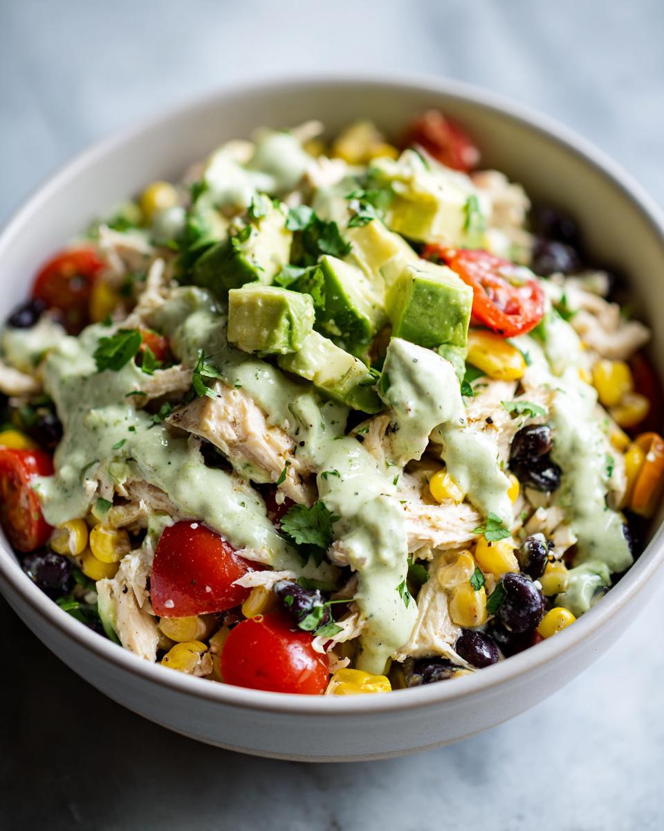 A bowl of vibrant southwest chicken salad topped with avocado chunks, black beans, corn, tomatoes, and creamy green dressing.