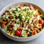 A vibrant bowl of southwest chicken salad featuring shredded seasoned chicken, corn, black beans, tomatoes, and topped with avocado chunks and creamy green dressing.