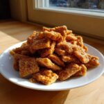 A pile of crunchy, golden-brown toffee chex mix recipe pieces coated in sparkling sugar on a white plate.