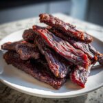 A pile of dark, richly seasoned venison jerky strips stacked on a white plate.