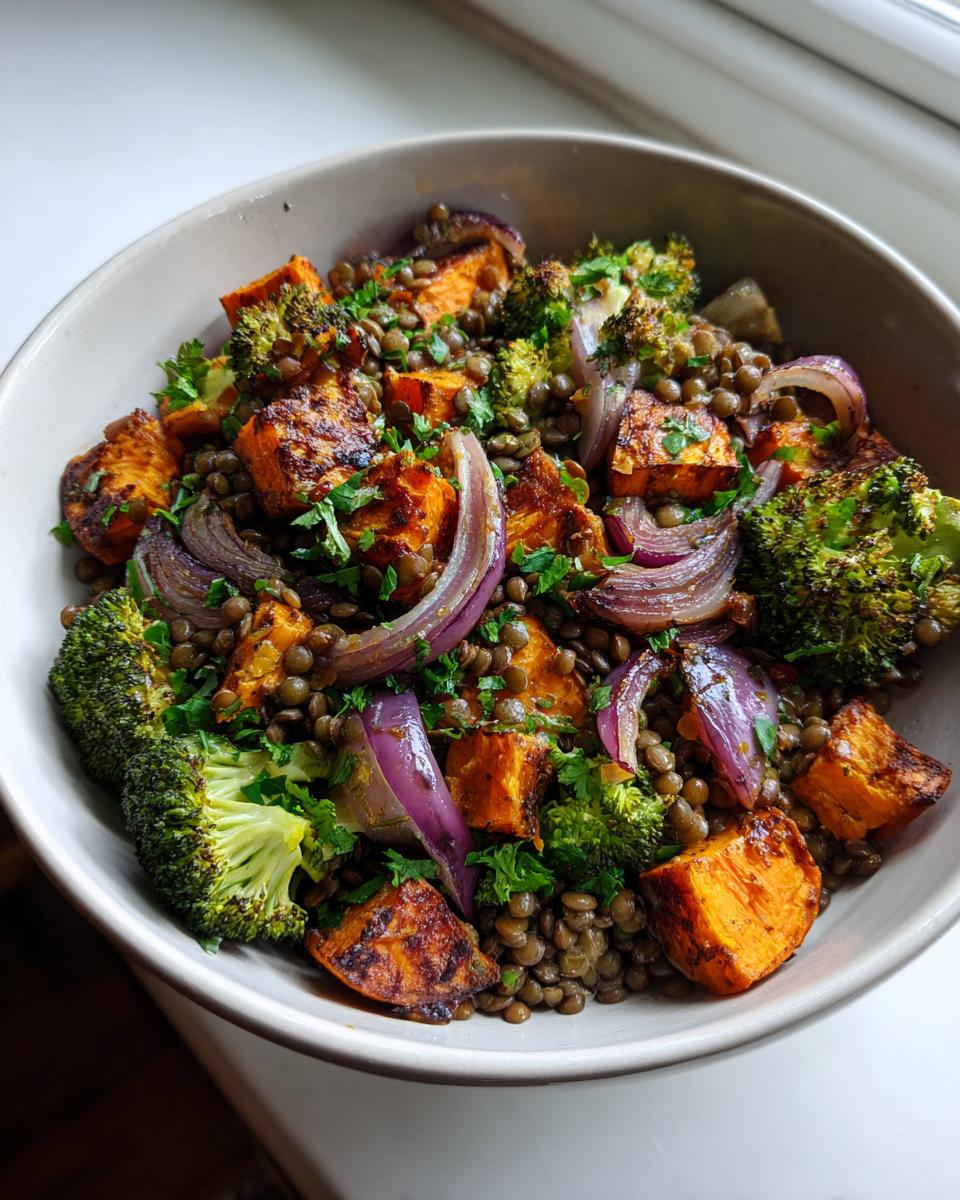 A close-up of a bowl filled with veggie lentil salad featuring green lentils, roasted sweet potatoes, purple onions, and broccoli florets.