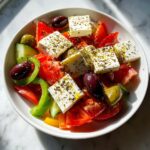 Close-up of a fresh greek salad featuring large cubes of feta cheese topped with oregano, tomatoes, cucumbers, peppers, and Kalamata olives.