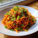 A close-up of a vibrant raw carrot salad, featuring spiralized carrots topped with green onions and sesame seeds.