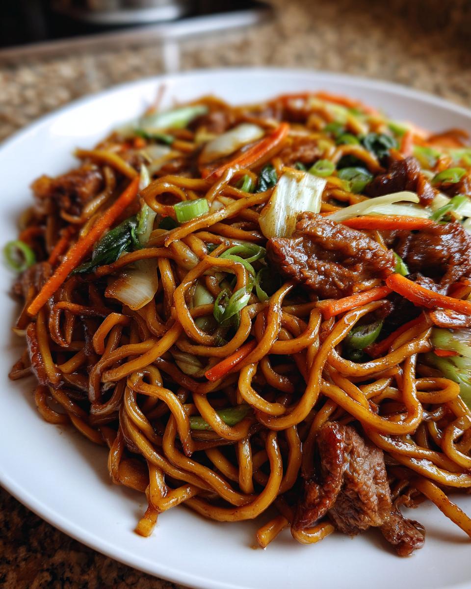 A close-up shot of a plate piled high with glossy, savory fried noodles mixed with beef and colorful vegetables.