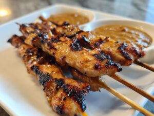 Close-up of grilled chicken satay skewers with visible char marks, served on a white plate with peanut dipping sauce.