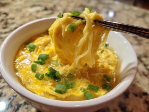 Chopsticks lifting ribbons of cooked egg from a bowl of savory egg drop soup garnished with green onions.