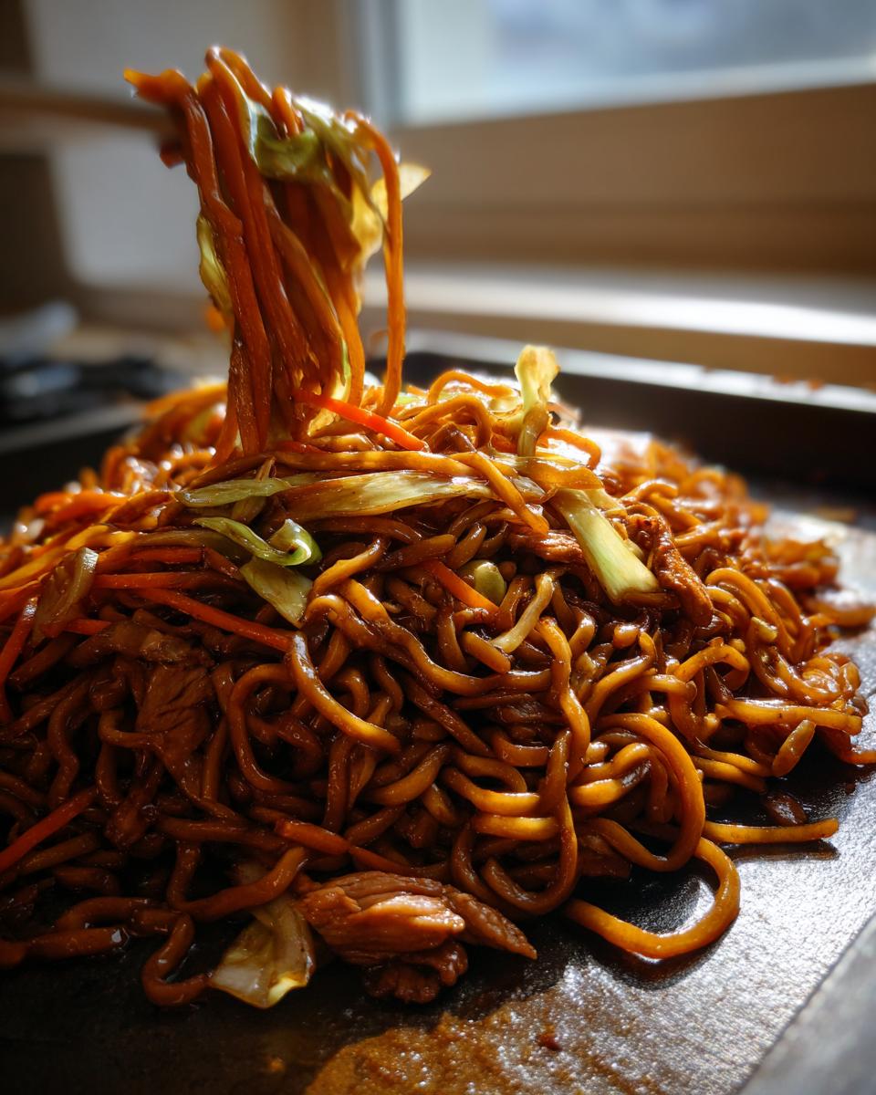 A close-up shot showing a large serving of dark, saucy fried noodles being lifted by chopsticks from a hot griddle.