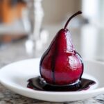 A single, deep magenta poached pear drizzled with its dark syrup, resting on a white plate.