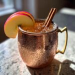 A frosty copper mug filled with an apple cider Moscow mule, garnished with apple slices and cinnamon sticks.