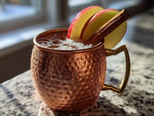 A frosty copper mug filled with an apple cider moscow mule, garnished with apple slices and a cinnamon stick.