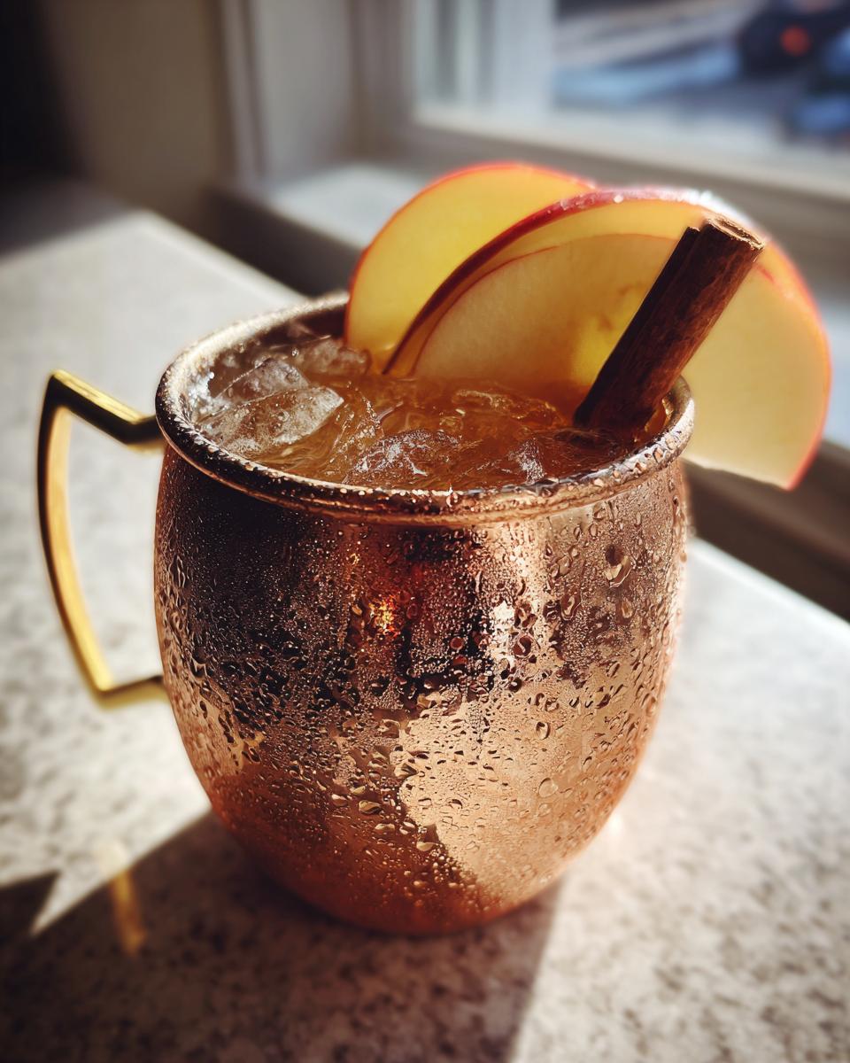 A refreshing apple cider moscow mule served over ice in a frosty copper mug, garnished with apple slices and a cinnamon stick.