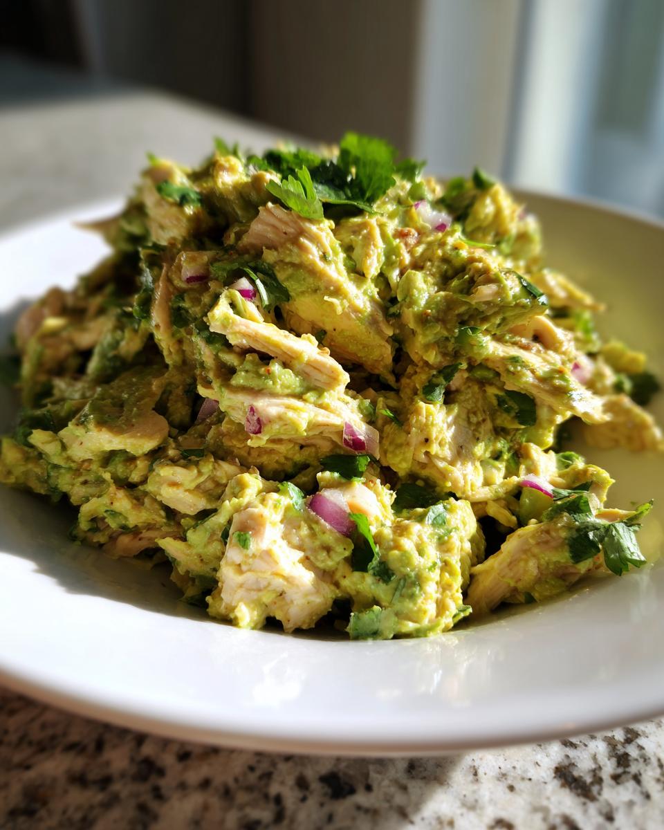 A close-up of creamy avocado chicken salad mixed with red onion and cilantro, served on a white plate.