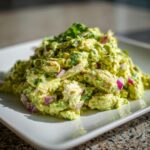 Close-up of a mound of creamy avocado chicken salad mixed with red onion and cilantro on a white plate.