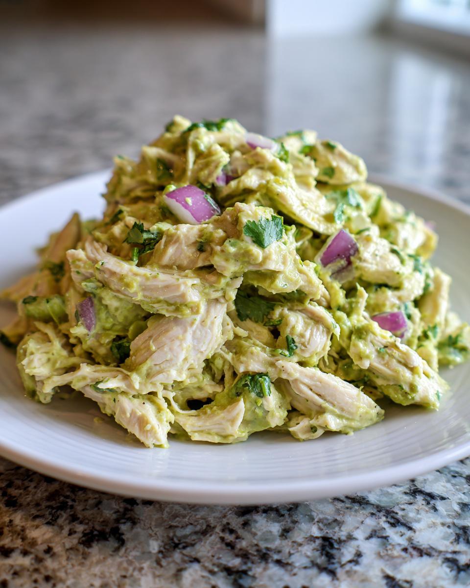 A mound of creamy avocado chicken salad mixed with chunks of red onion and cilantro, served on a white plate.