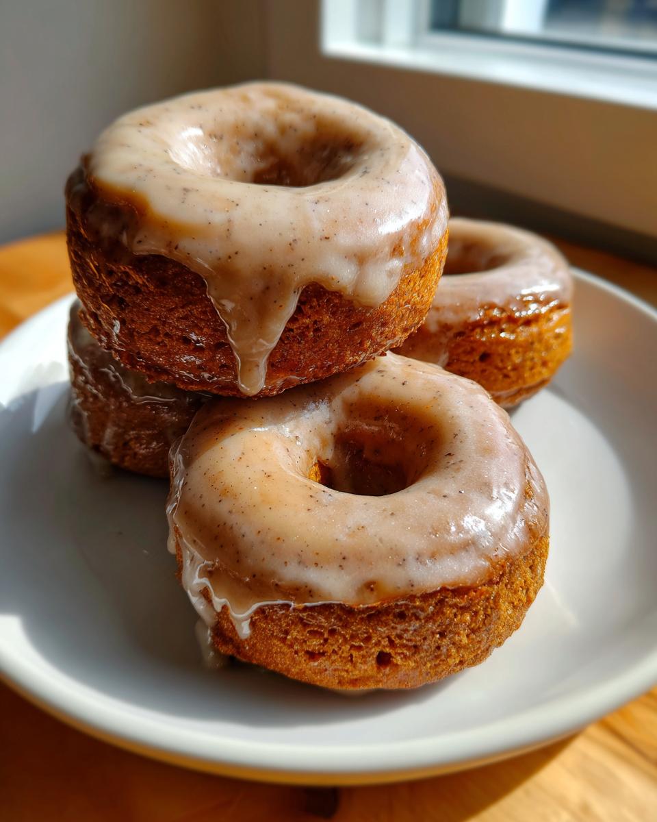 A stack of three golden-brown baked pumpkin donuts topped with a speckled vanilla glaze, sitting on a white plate.