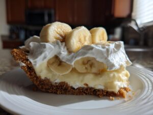 A generous slice of banana cream pie recipe featuring a graham cracker crust, thick vanilla custard, and topped with whipped cream and fresh banana slices.