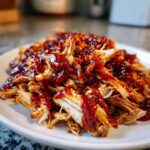 Close-up of tender, shredded pulled chicken generously coated in glossy barbecue sauce on a white plate.