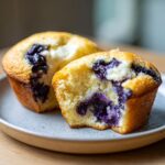 Two moist blueberry cream cheese muffins, one broken open to show the filling, resting on a speckled plate.