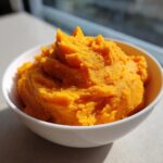 A close-up of vibrant orange mashed sweet potatoes served in a white bowl, catching bright sunlight.