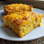 Two golden slices of cheesy cornbread casserole featuring visible corn kernels, served on a white plate.