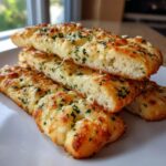 Close-up of several golden brown, cheesy garlic breadsticks topped with chopped parsley, stacked on a white plate.