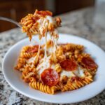 A fork lifting a bite of cheesy pizza pasta, showing long cheese pulls over a bowl of rotini pasta, sauce, and pepperoni.