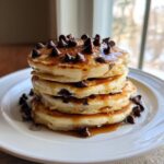 A tall stack of four fluffy chocolate chip pancakes drizzled with syrup and topped with chocolate chips.