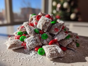 A mound of powdered sugar-coated christmas puppy chow mixed with red and green M&Ms, set near a window with a blurred Christmas tree.