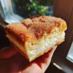 A hand holds a square slice of churro cheesecake topped with sparkling cinnamon sugar.