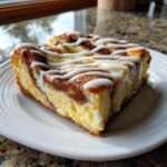 A close-up of a rich slice of cinnamon roll cheesecake drizzled with white icing.