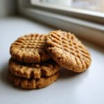 A small stack of four golden brown peanut butter cookies showing classic crosshatch fork marks, resting on a white surface.