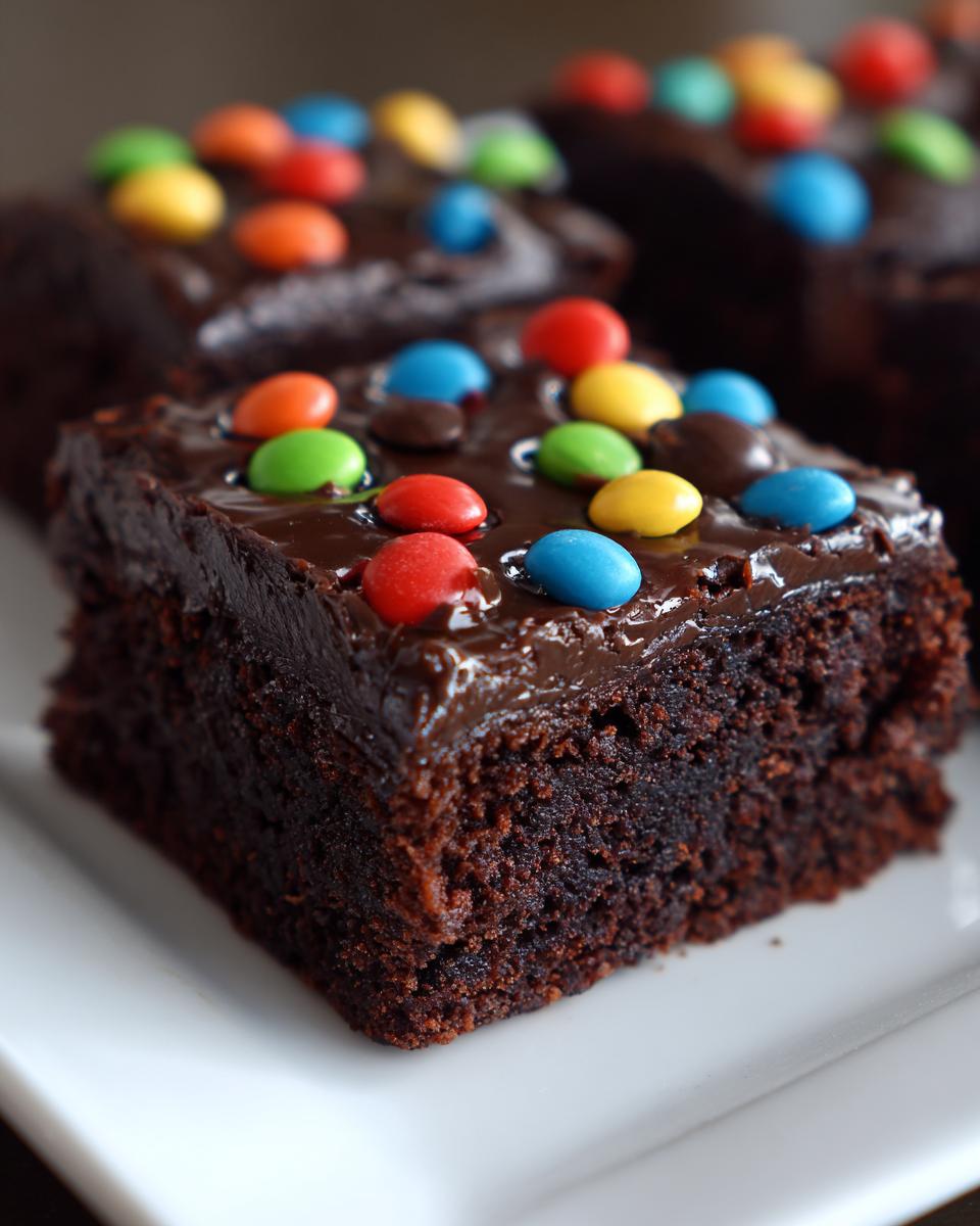 A close-up of a fudgy square cosmic brownies slice topped with glossy chocolate frosting and colorful candy pieces.