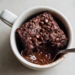 A close-up of a rich, dark Cottage Cheese Microwave Brownie served in a white mug with a spoonful revealing a gooey center.