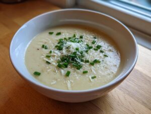 A close-up of a bowl of creamy cauliflower soup garnished with grated cheese and fresh chopped chives.