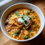 A close-up of a hearty bowl of chicken tortilla soup, topped with sour cream, tortilla strips, and cilantro.