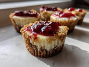 A close-up, sunlit photo of a single mini cheesecake with a graham cracker crust and bright red cherry topping, with more mini cheesecake bites blurred in the background.